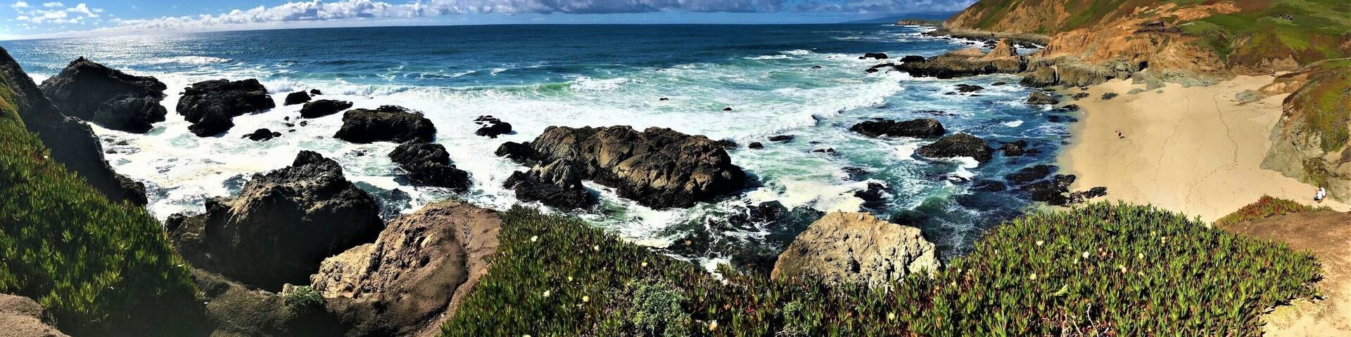 A coastal landscape with rocky shores, a sandy beach, and green hills under a clear blue sky.
