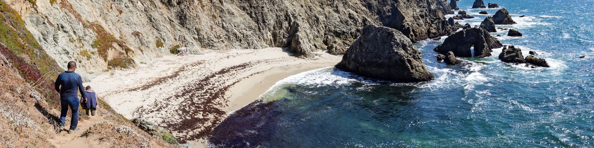 A coastal landscape with a steep cliff, a sandy beach, and large boulders in the water.