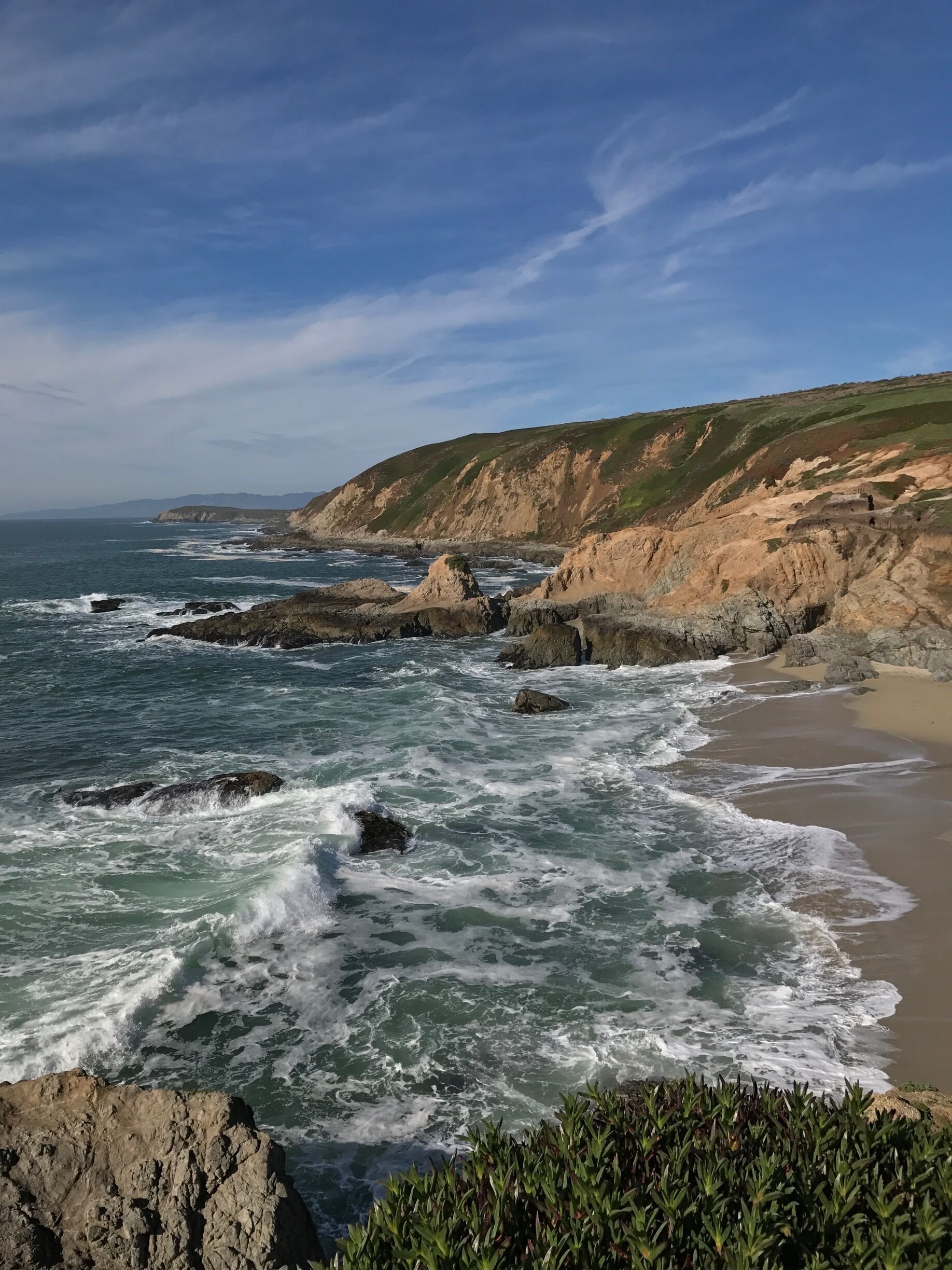 A coastal landscape with rocky cliffs, waves crashing onto the shore, and a clear sky.