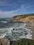 A coastal landscape with rocky cliffs, waves crashing onto the shore, and a clear sky.