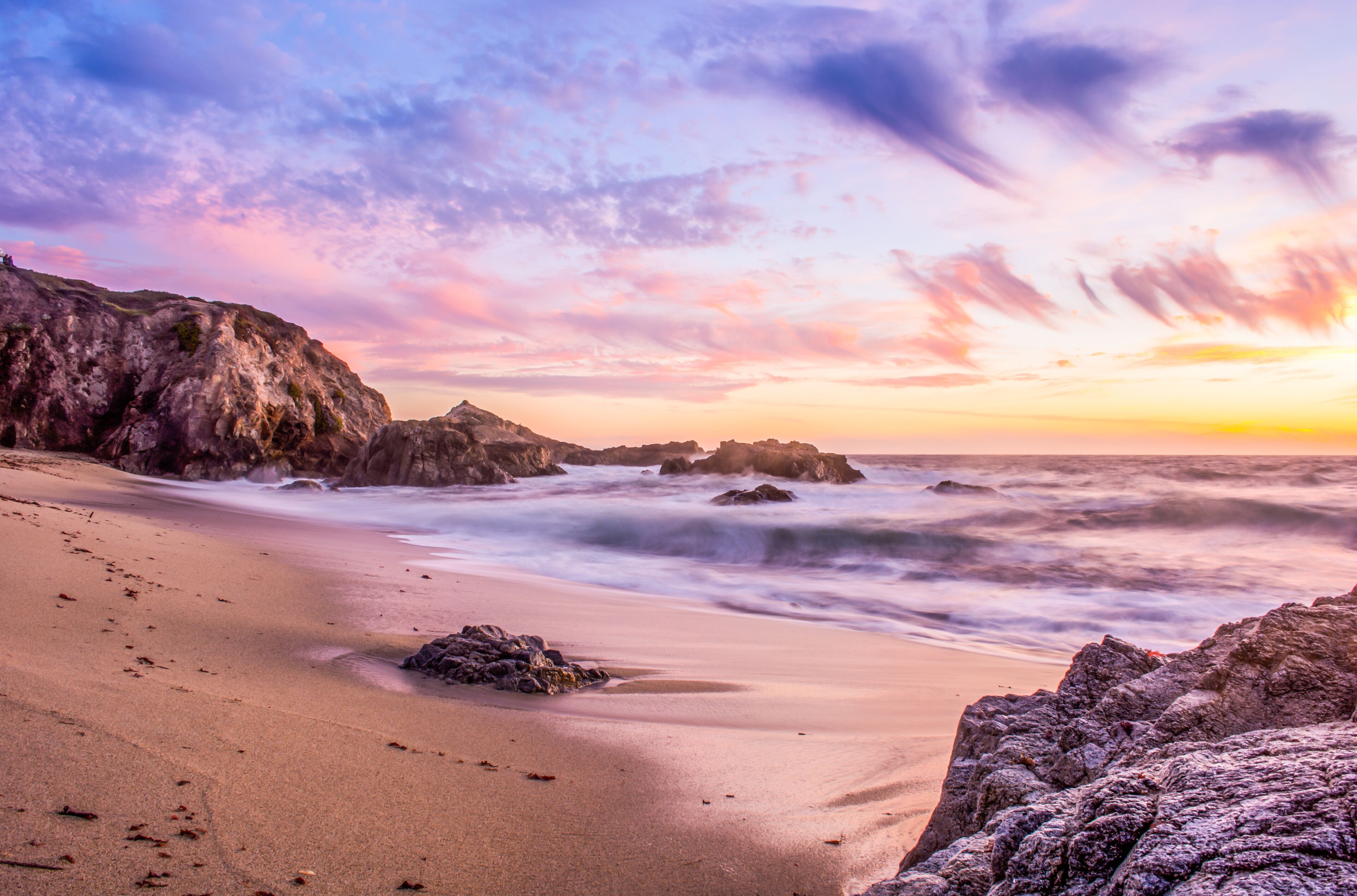 A beach with smooth sand, rocky outcrops, and a sunset sky.