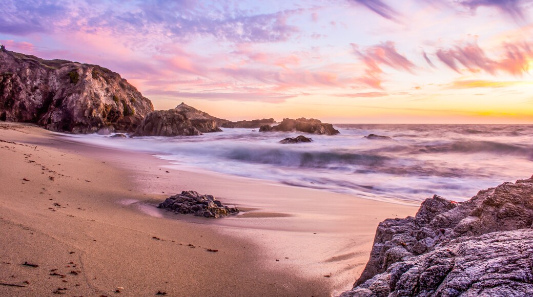 A beach with smooth sand, rocky outcrops, and a sunset sky.
