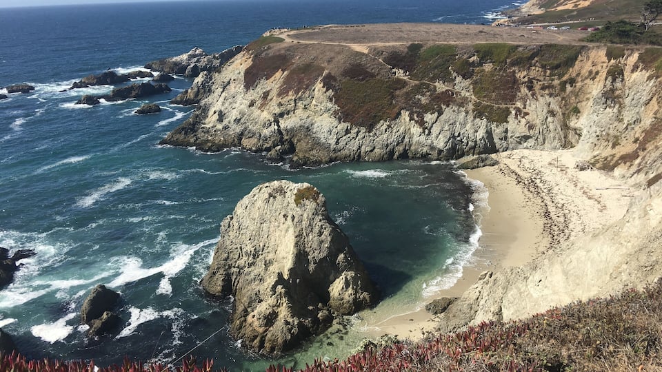 A coastal landscape with rocky cliffs, a sandy beach, and clear blue ocean waves.