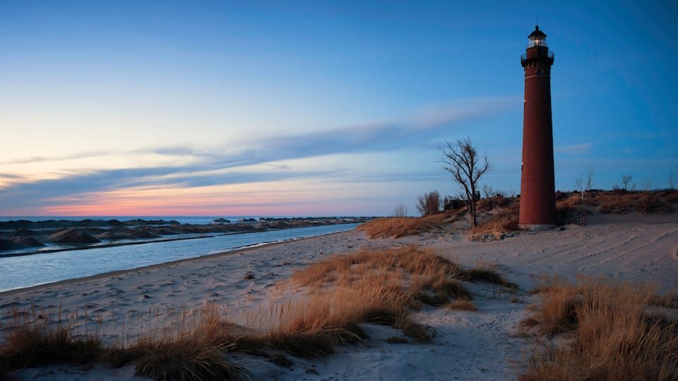 A red lighthouse on a sandy beach with sparse grass and a clear sky.