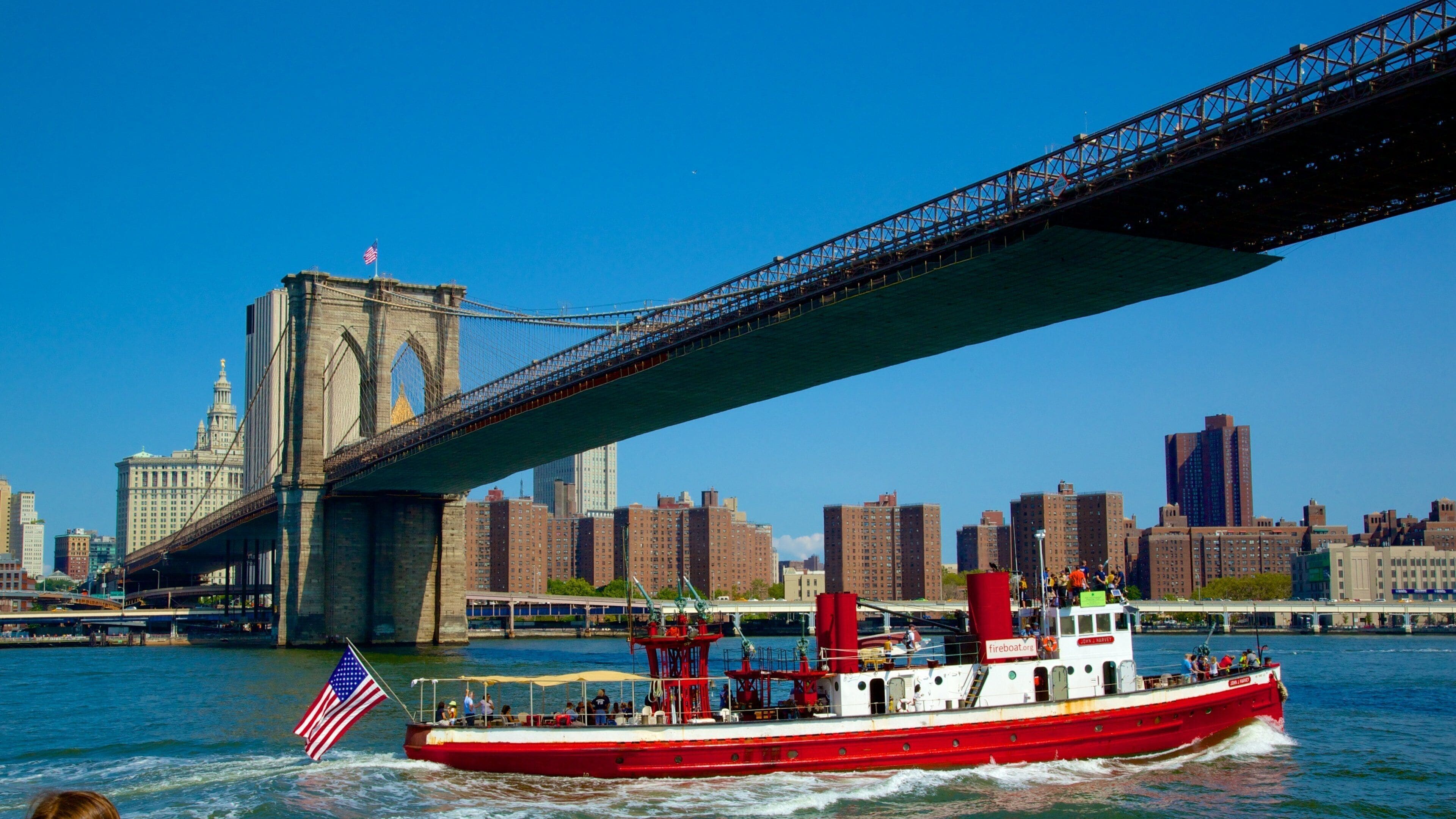 Un vaixell vermell amb bandera estatunidenca al riu Hudson, passant sota el Pont de Brooklyn amb l'horitzó de Manhattan al fons.