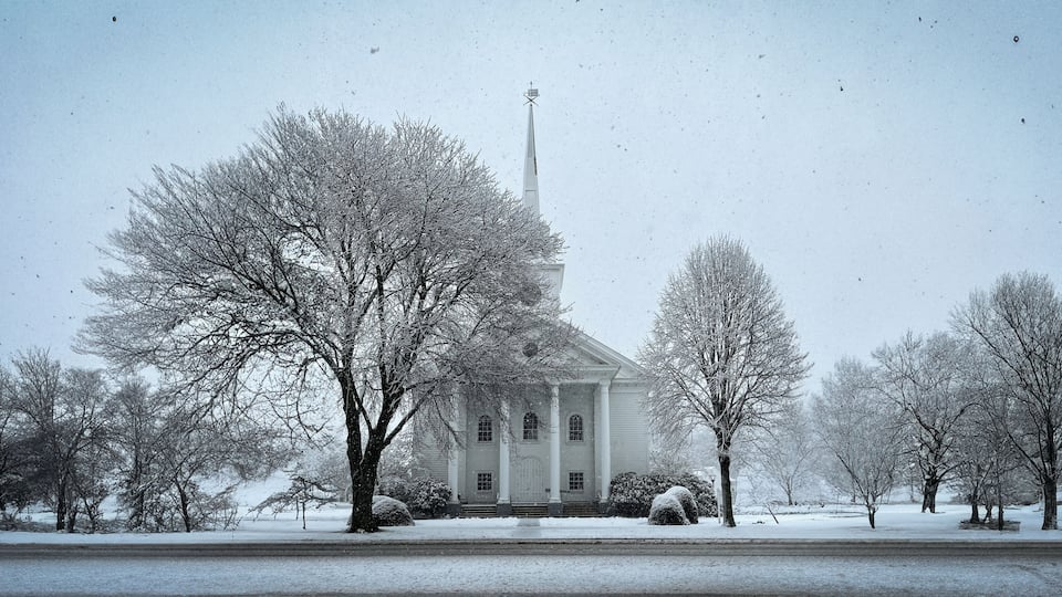 A snow-covered church with a steeple, surrounded by leafless trees and a snow-covered lawn.
