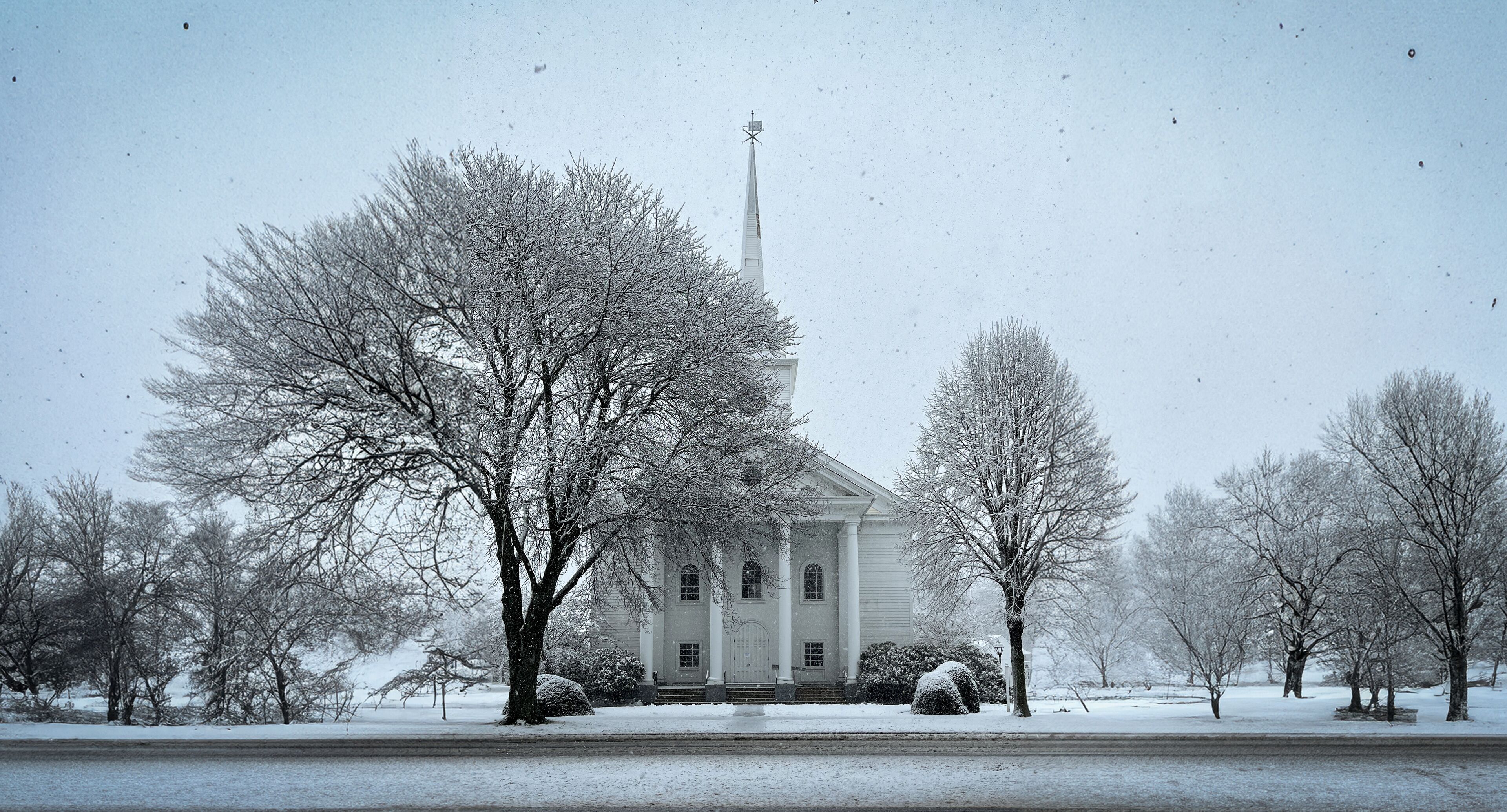 A snow-covered church with a steeple, surrounded by leafless trees and a snow-covered lawn.