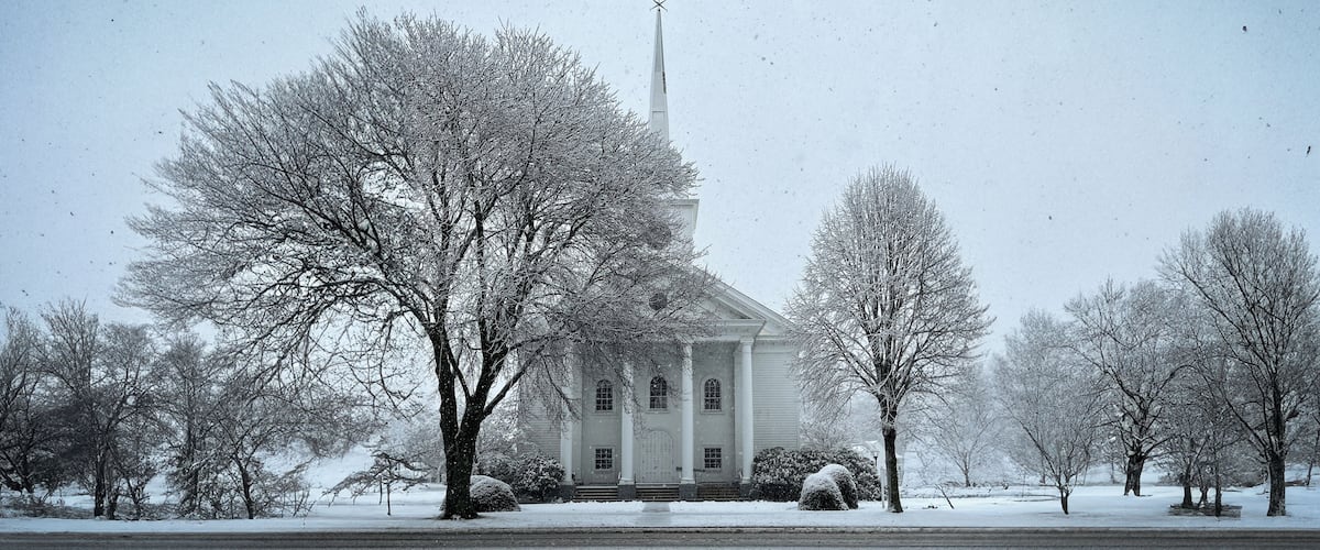 A snow-covered church with a steeple, surrounded by leafless trees and a snow-covered lawn.