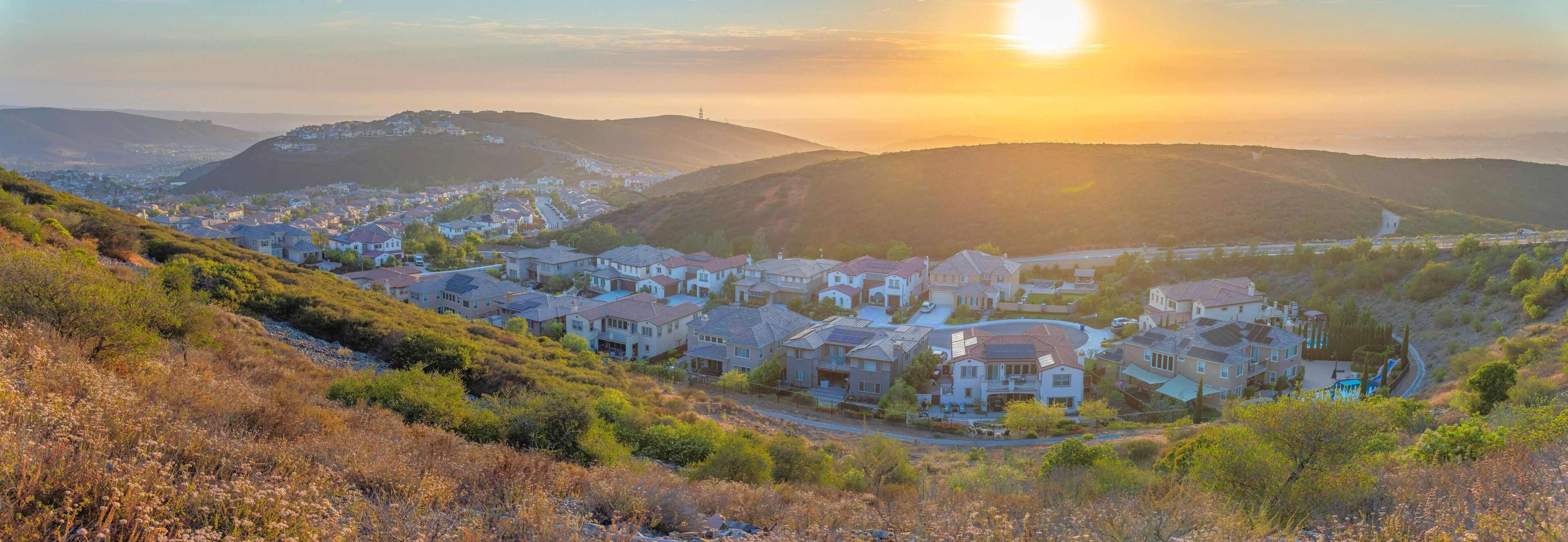 A residential area with houses on a hillside, a clear sky, and the sun setting in the background.