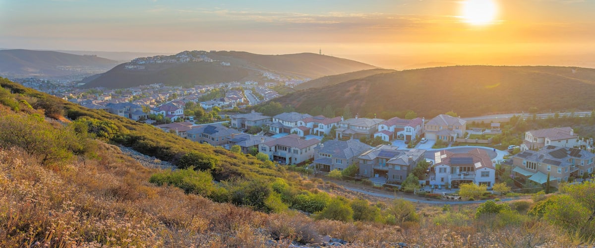 A residential area with houses on a hillside, a clear sky, and the sun setting in the background.