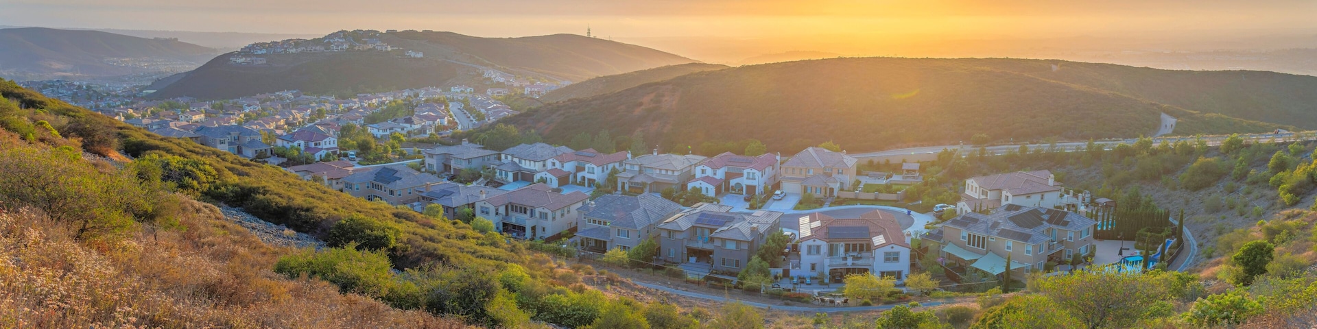 A residential area with houses on a hillside, a clear sky, and the sun setting in the background.