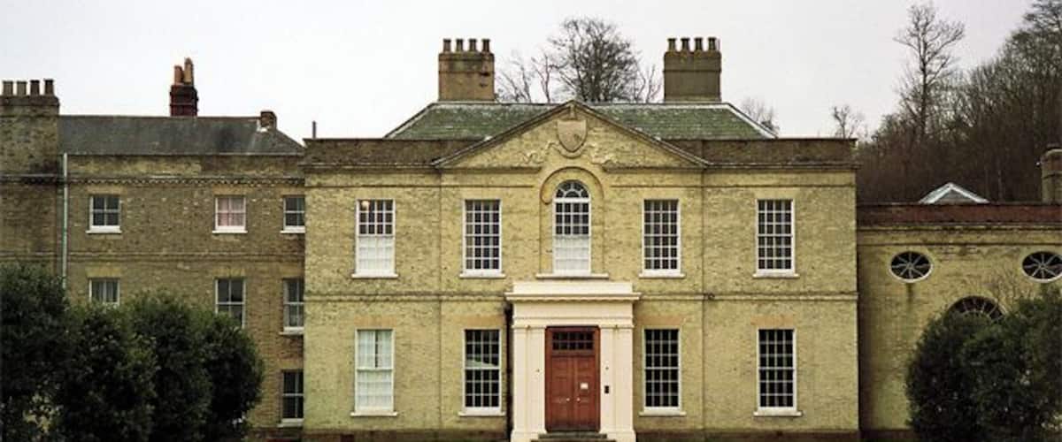 A large, historic building with a central entrance, symmetrical windows, and a gravel driveway.