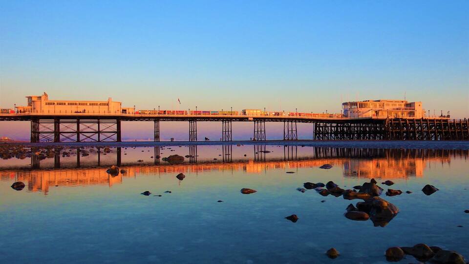 A pier extending into calm waters with rocks in the foreground and a clear sky.