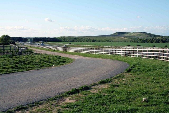 Un camino rural serpentea a través de un paisaje cubierto de hierba, con una valla blanca y colinas lejanas.