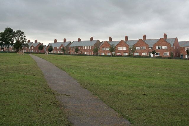 A row of red brick houses with a paved path leading through a grassy field.