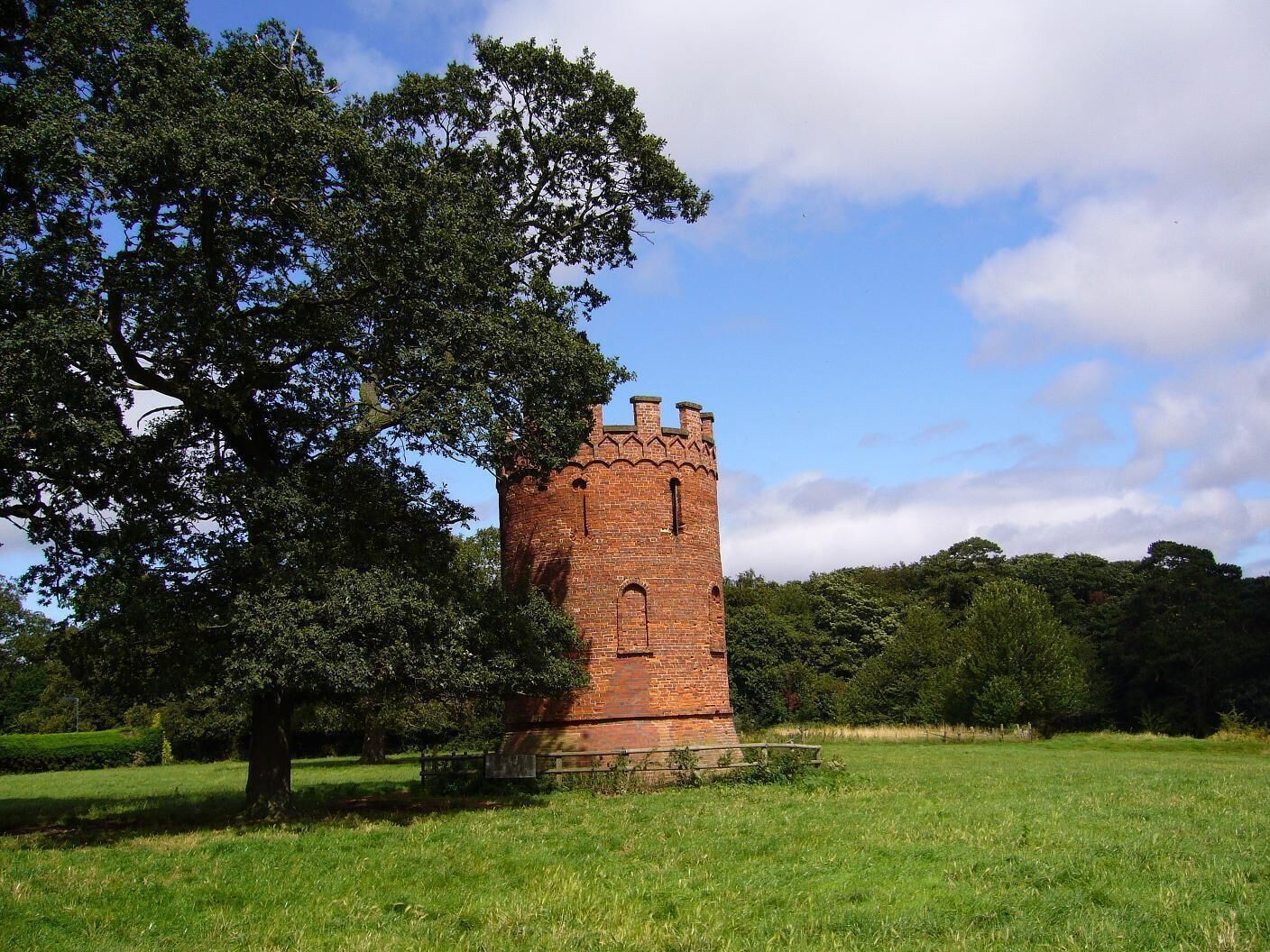 A brick tower in a grassy field with a large tree nearby.