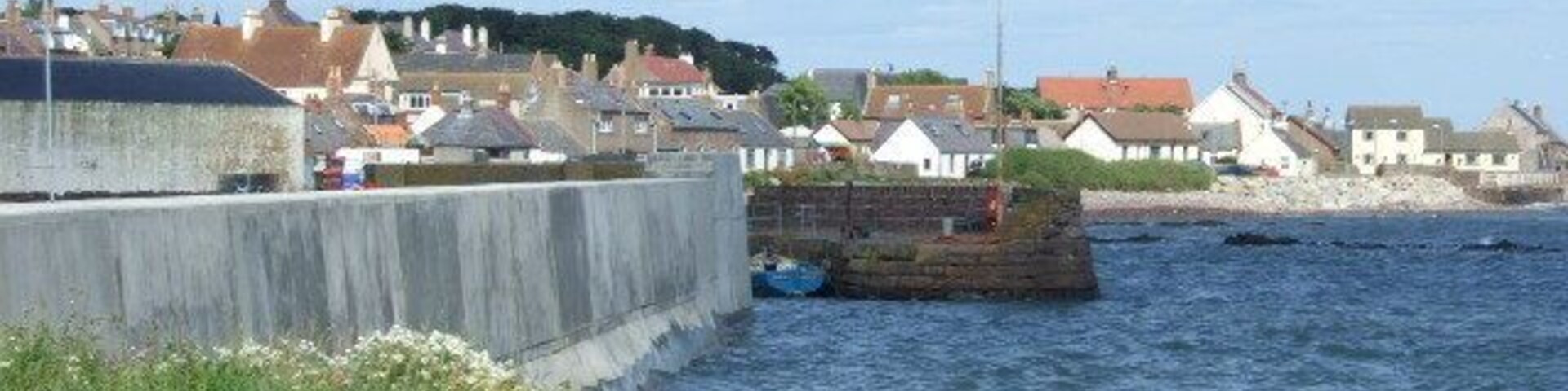 Uma vila costeira com um muro de pedra à beira-mar, casas e um afloramento rochoso na água.