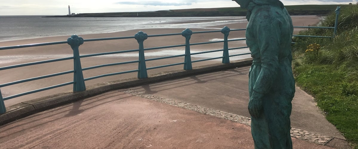 A statue on a coastal walkway with a view of the sea and a lighthouse in the distance.