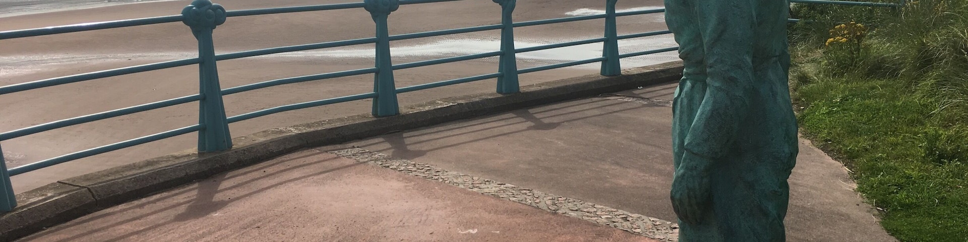 A statue on a coastal walkway with a view of the sea and a lighthouse in the distance.