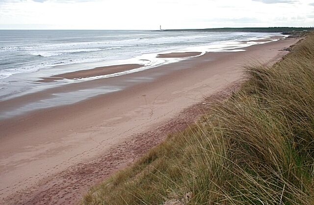 A sandy beach with waves, grassy dunes, and a distant lighthouse.