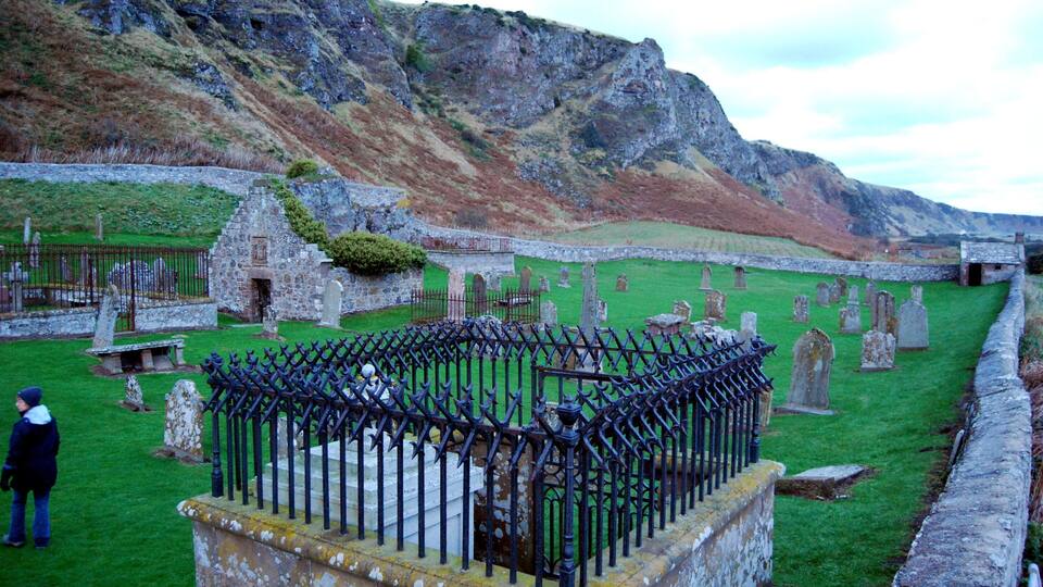A cemetery with stone headstones and a stone building, surrounded by grassy areas and a rocky hill.