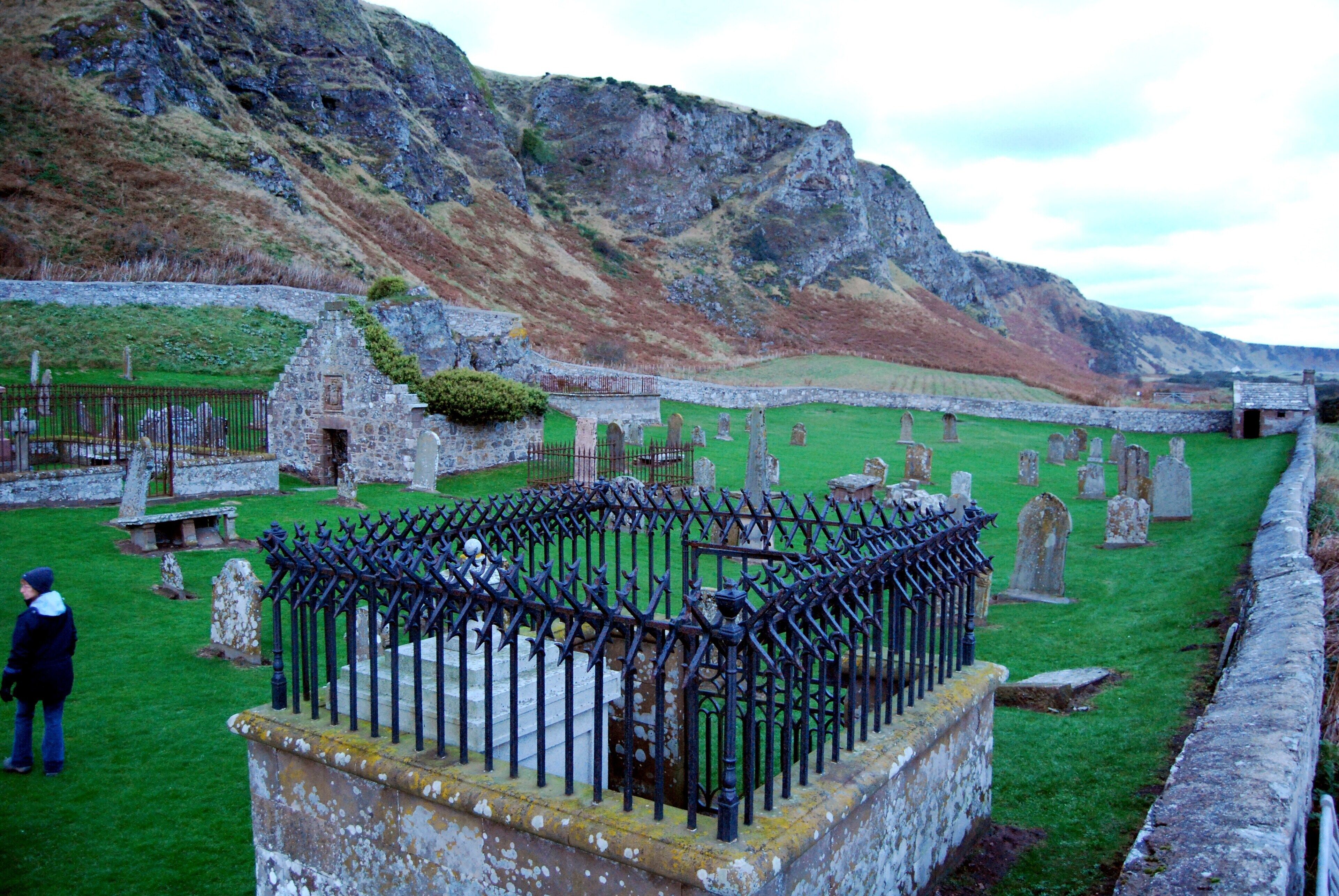 A cemetery with stone headstones and a stone building, surrounded by grassy areas and a rocky hill.