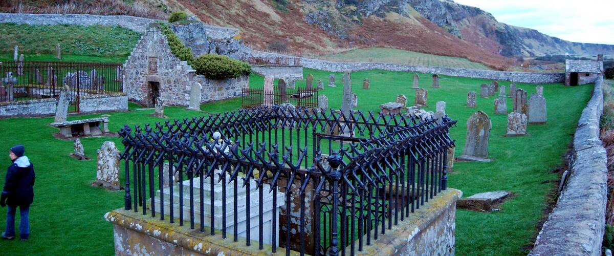 A cemetery with stone headstones and a stone building, surrounded by grassy areas and a rocky hill.
