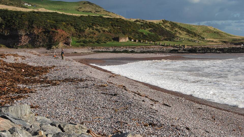 A pebbly beach with waves, a rocky shoreline, and a grassy hill with a small building in the distance.