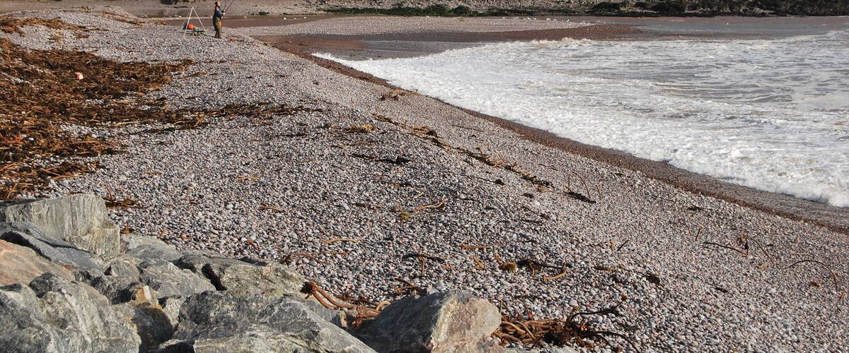 A pebbly beach with waves, a rocky shoreline, and a grassy hill with a small building in the distance.