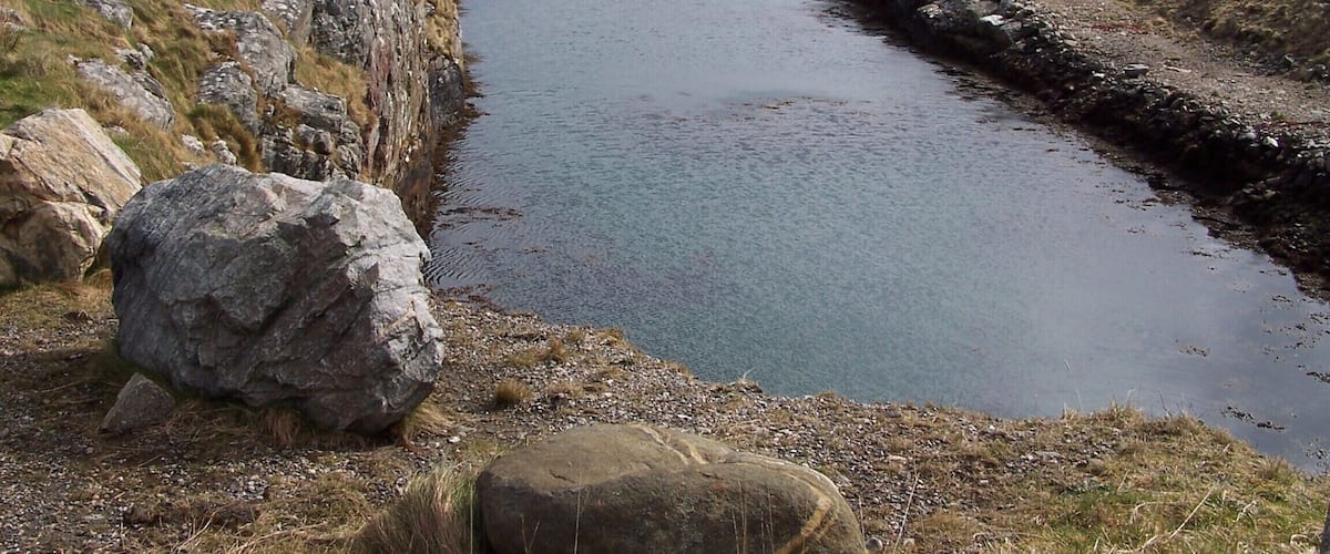 A coastal landscape with a narrow waterway, rocky shores, and distant hills.