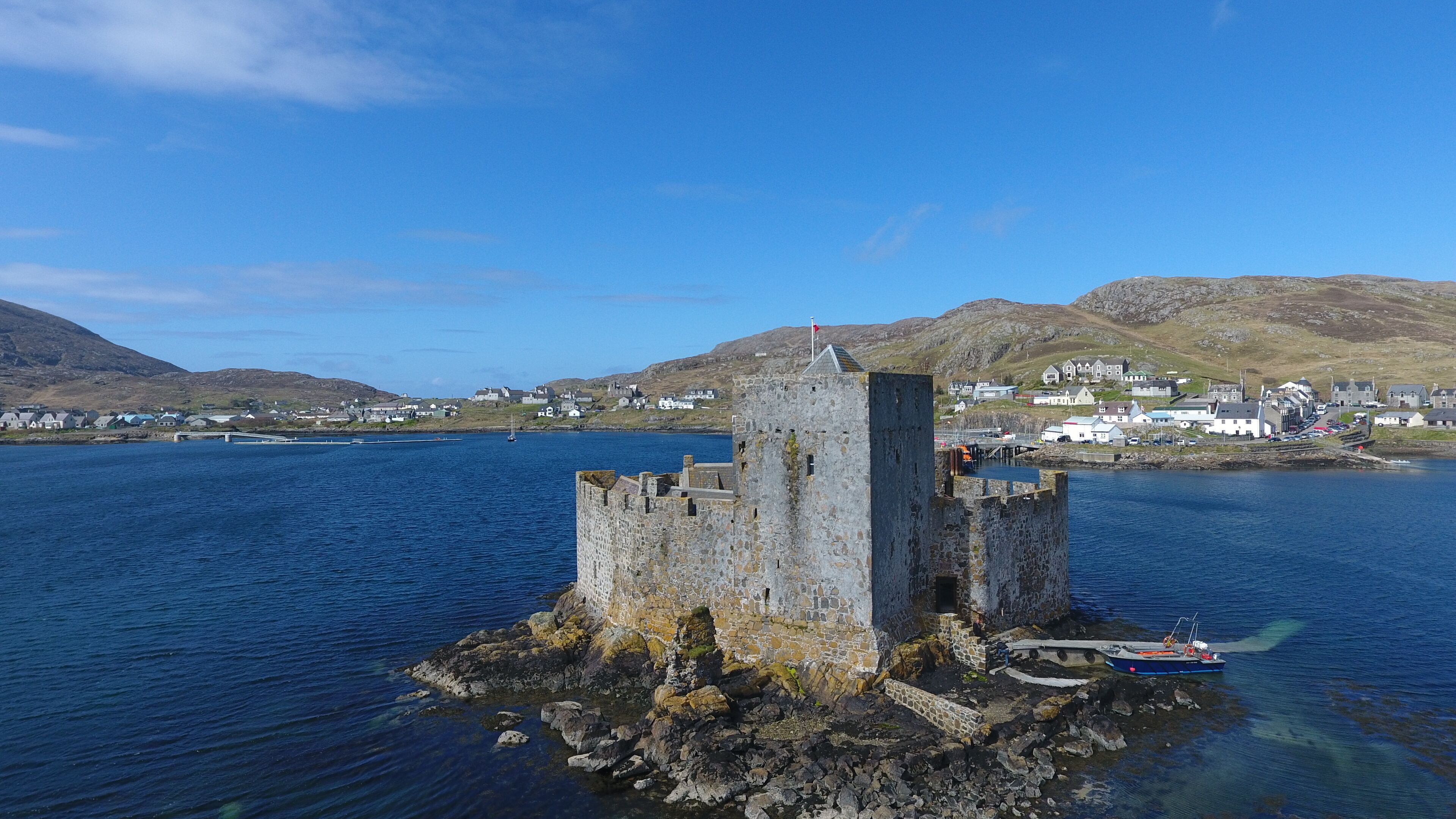 A stone tower surrounded by water, with a small boat nearby. In the background, there are houses and hills.