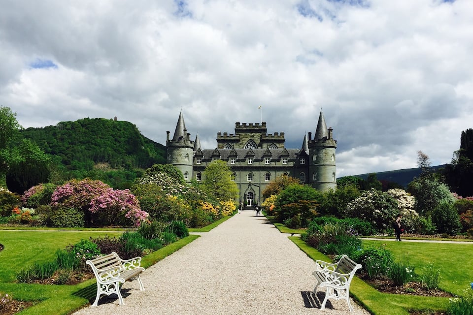 Un castillo con un camino de grava que conduce a él, rodeado de jardines exuberantes y árboles.