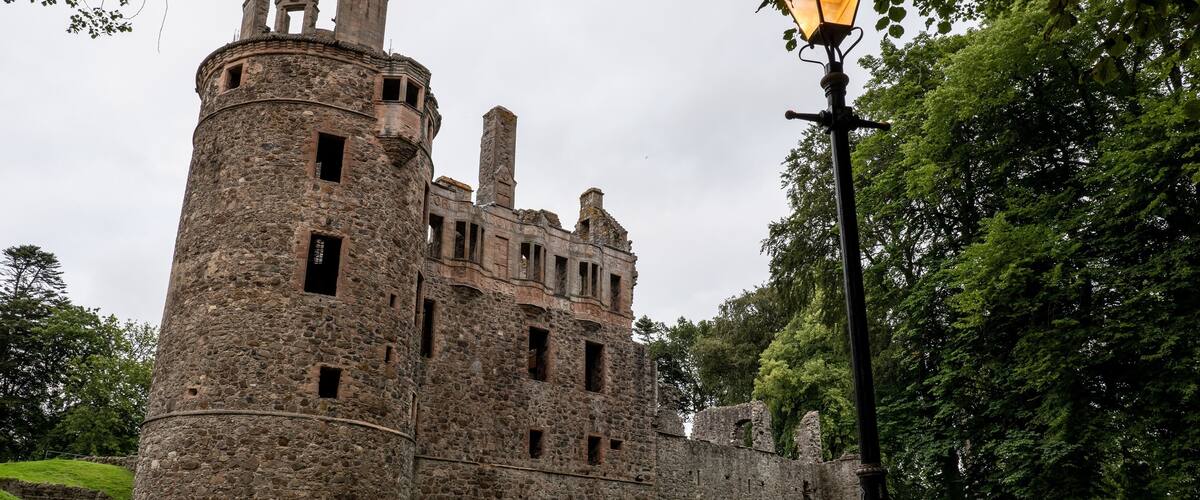 A historic stone castle with a round tower, surrounded by lush greenery and a street lamp.