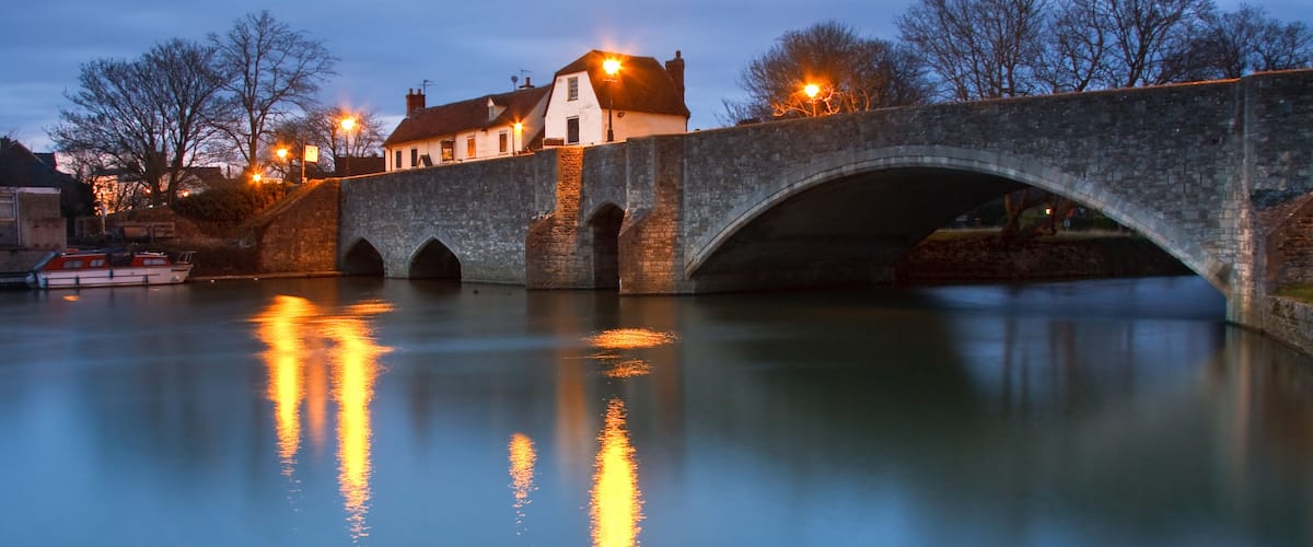 A stone bridge over a calm river, with buildings and street lamps in the background.