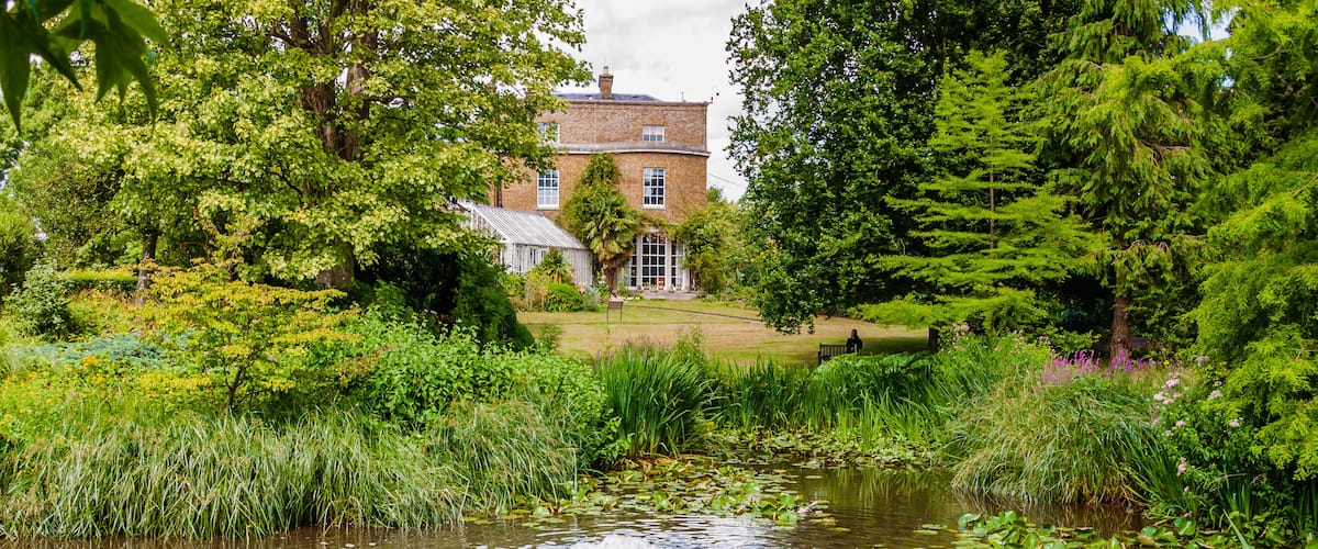 A pond surrounded by lush greenery and a brick building in the background.
