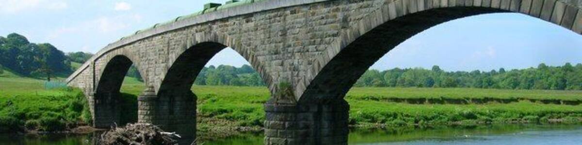 Eine Steinbrücke über einem ruhigen Fluss, im Hintergrund grüne Felder und Bäume.