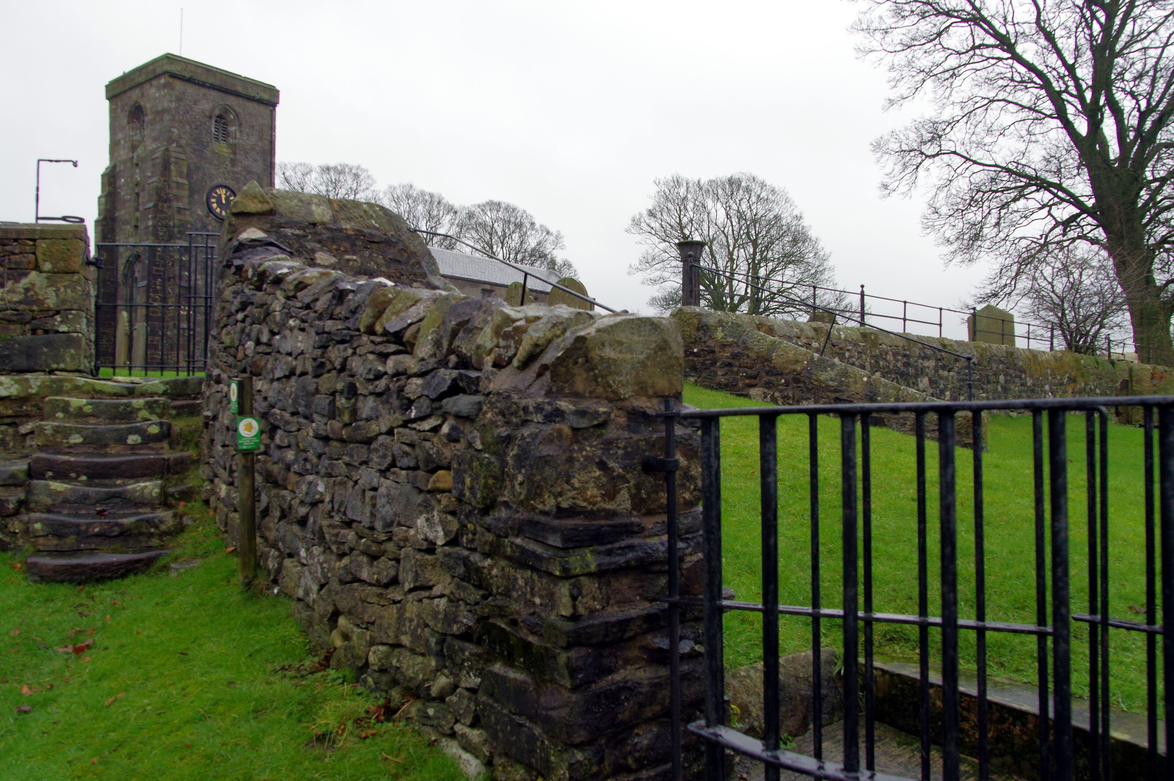 A stone wall with a clock tower, a metal fence, and a grassy area.