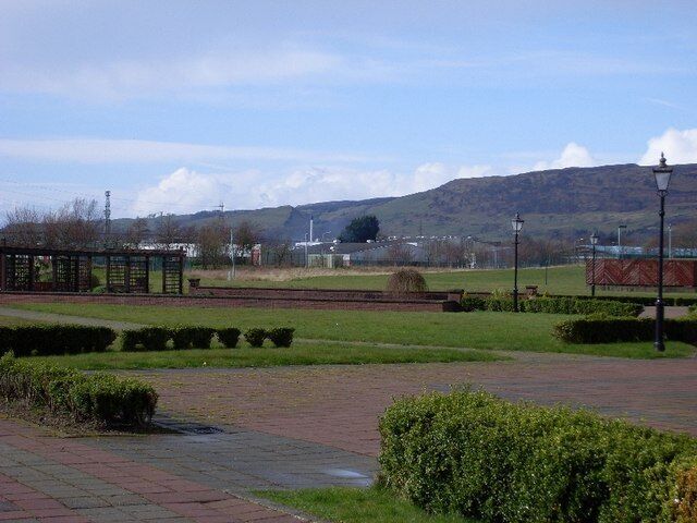 Um parque com calçada pavimentada, gramados verdes, uma construção pequena e colinas ao longe.