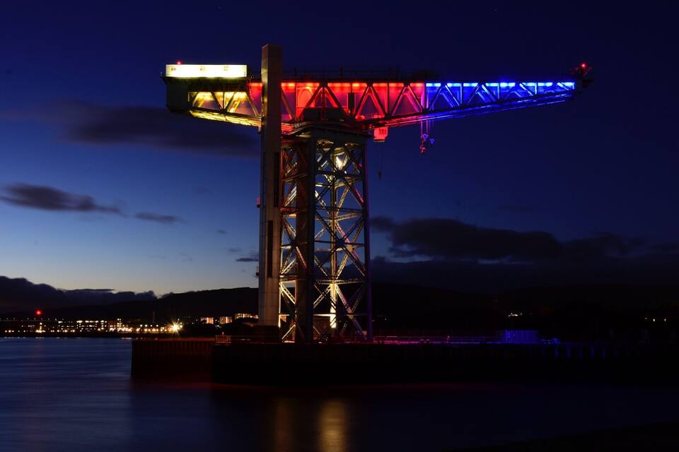 A lit-up crane structure at night with a cityscape in the background.