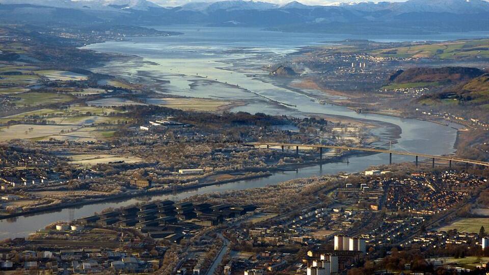 A river flowing through a town with a bridge, surrounded by mountains and fields.