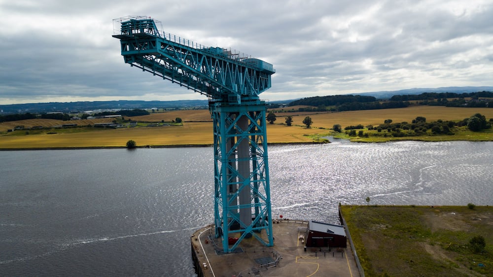 A large blue crane structure over a river with fields and hills in the background.