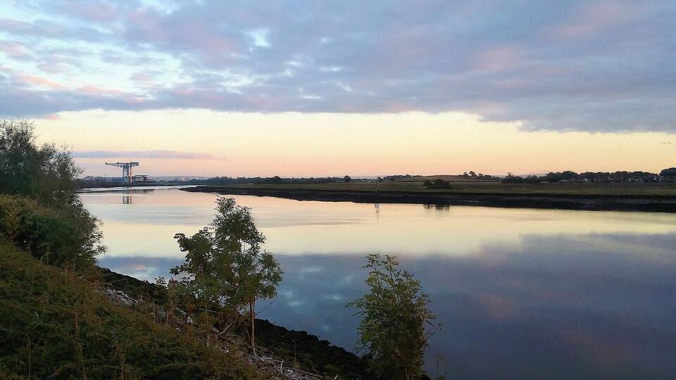 A calm river with a bridge in the distance, surrounded by trees and open fields.