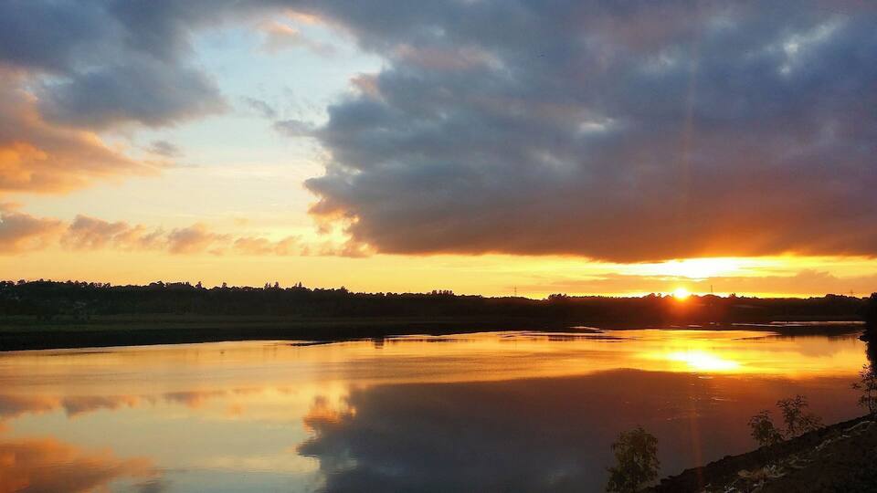 A sunset over a calm river with a silhouette of trees and a cloudy sky.