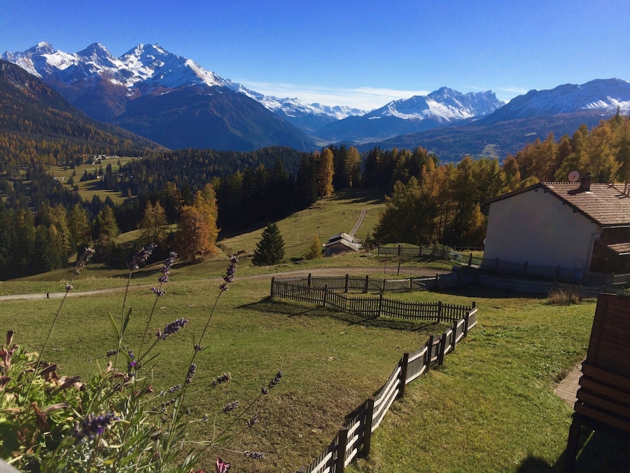 A mountainous landscape with snow-capped peaks, a wooden fence, and a small building.