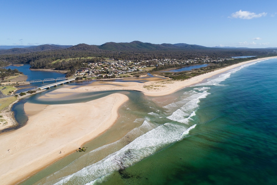A coastal landscape with a sandy beach, a clear blue ocean, and a bridge connecting two land masses.