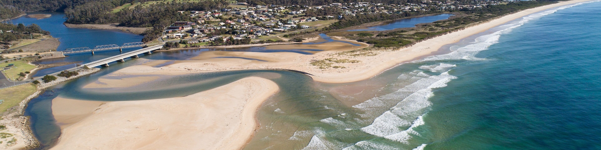 Un paisaje costero con una playa arenosa, un océano azul transparente y un puente que une dos extensiones de tierra.