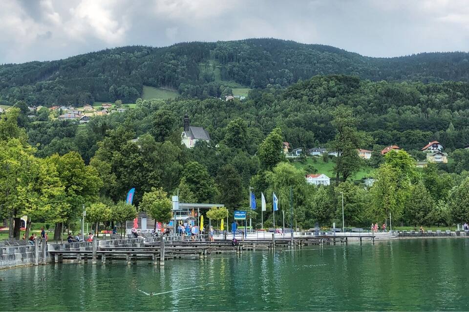 A lakeside park with a wooden pier, trees, and houses in the background.