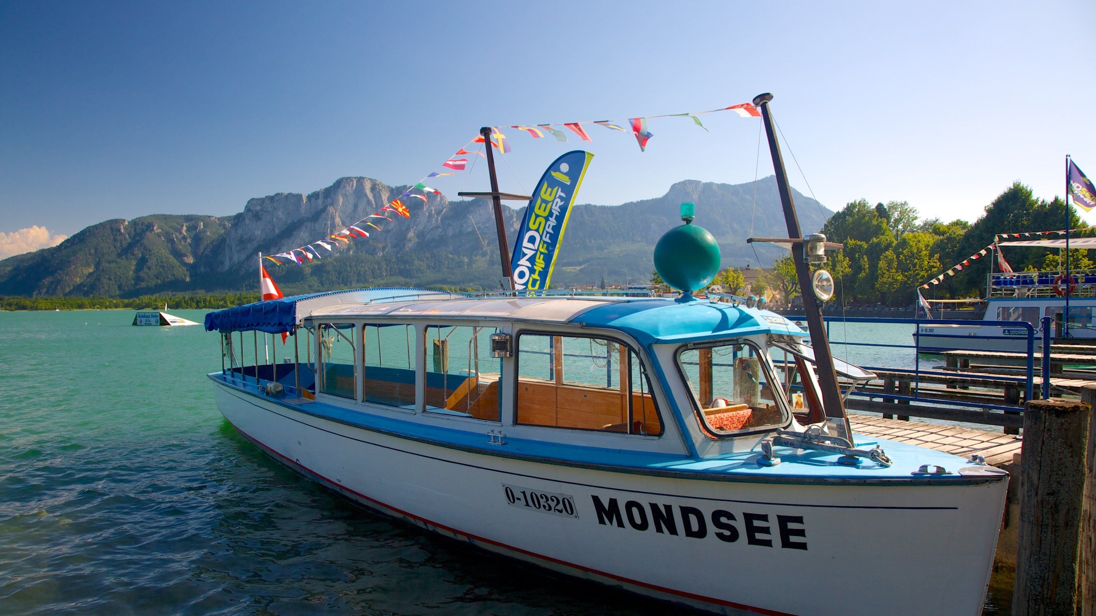 A boat named 'MONDSSEE' docked at a pier with mountains in the background.