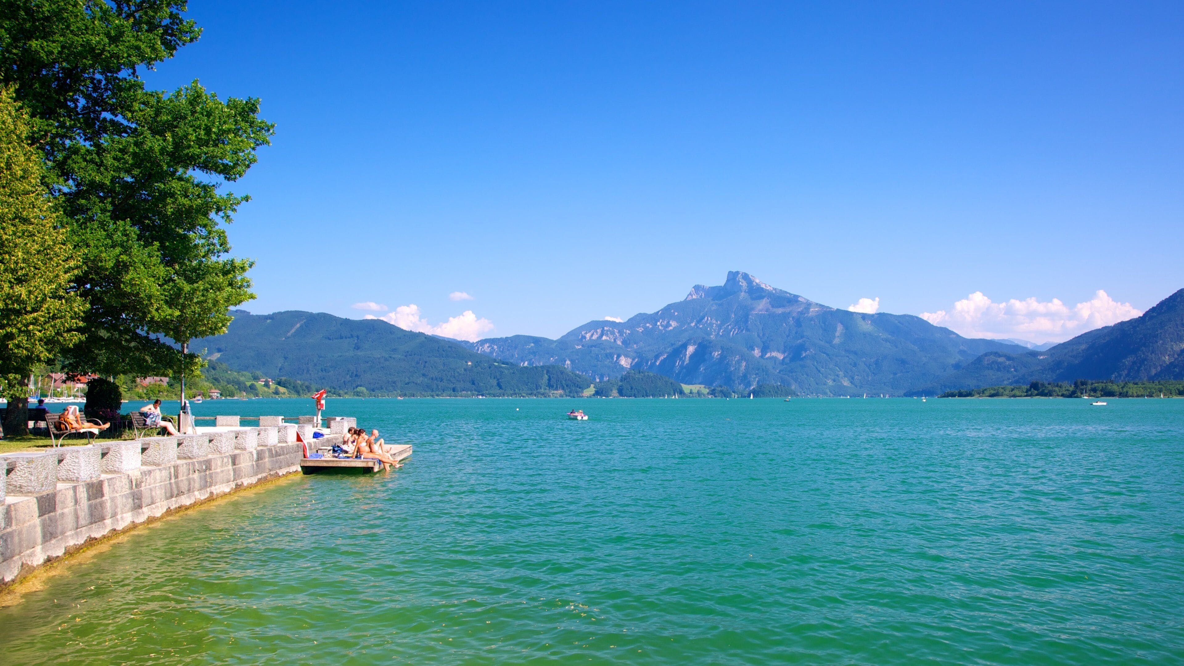 A lake with a boat, people relaxing on a dock, and mountains in the background.