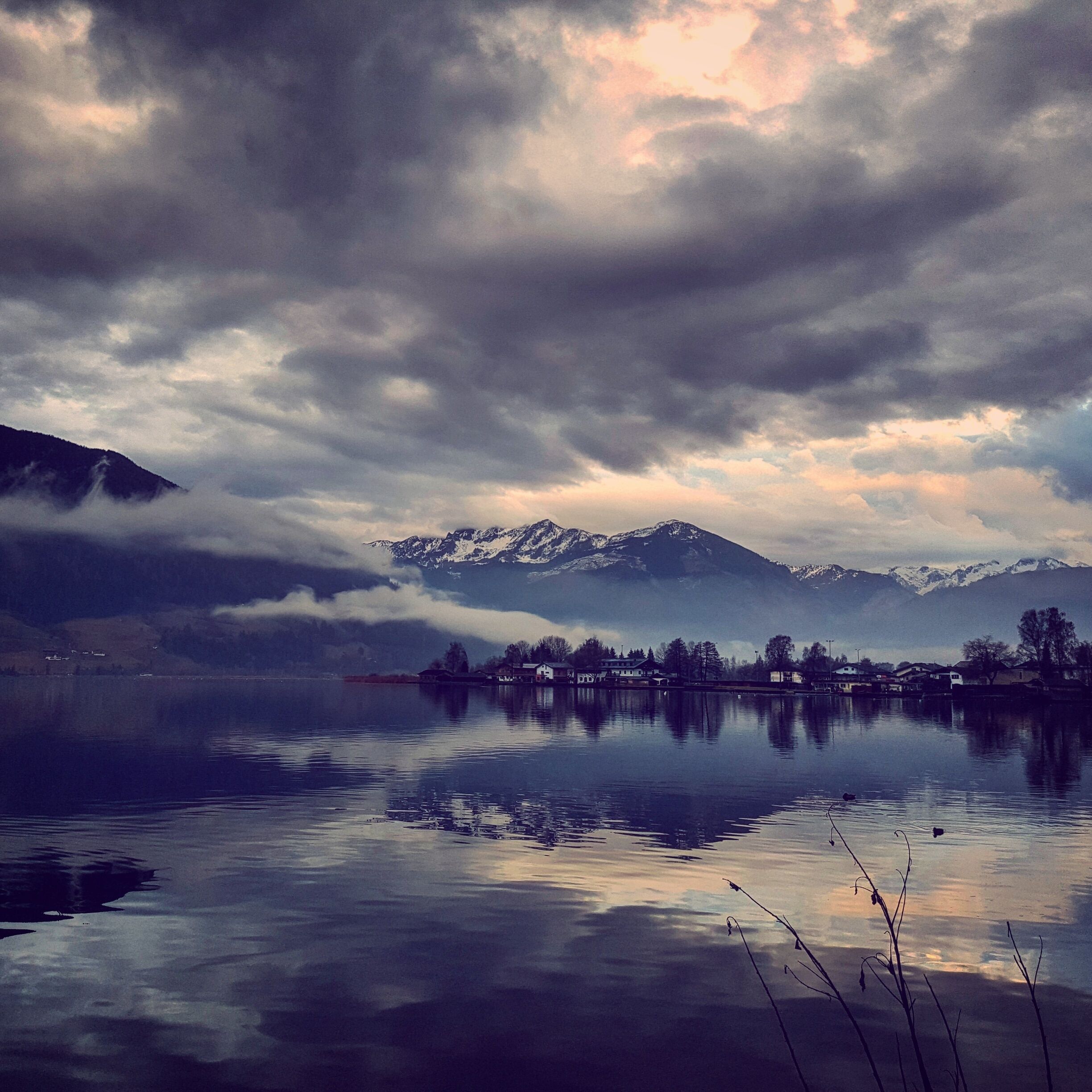 Un lago tranquilo con una cadena montañosa al fondo y un pequeño asentamiento al otro lado del agua.